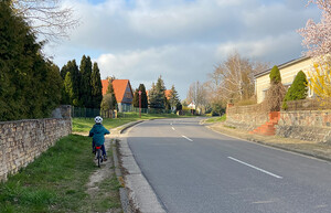 Landstraße in einem Ort. Kind fährt auf dem Trampelpfad neben der Straße, da es keinen Gehweg gibt.