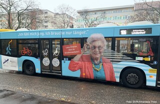 Ein Bus steht bei Regen an einer Haltestelle in Berlin. Auf dem Bus ist ein Bild mit einer älteren Frau. Sie trägt bunte Kleidung und neben ihr steht der Spruch: Lass Älteren Zeit im Straßenverkehr!