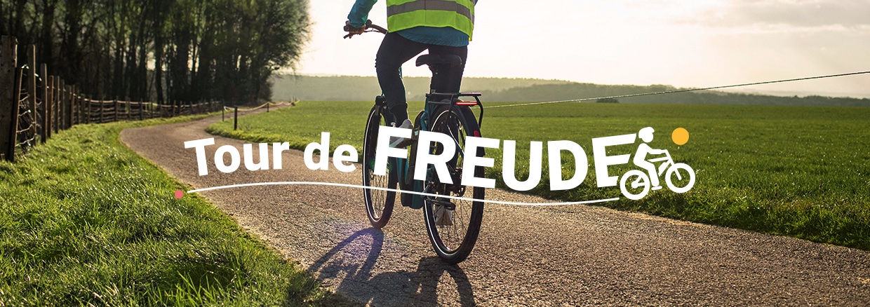 Kampagnenbanner Tour de Freude: Weiße Schrift vor einem Radfahrer auf einer Straße