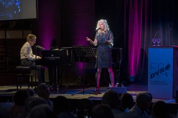 Jürgen Menge sitzt am Klavier und spielt. Neben ihm steht seine Frau Elli und singt. Die Beiden befinden sich auf einer Bühne. Neben der Frau ist ein Rednerpult zu sehen, woran ein Banner mit dem Spruch 50 Jahre DVR befestigt ist