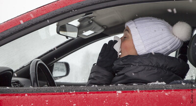 Eine Frau sitzt am Steuer eines roten Autos. Sie putzt sich die Nase, wobei ihre Augen geschlossen sind. Auf dem Kopf trägt sie eine weiße Mütze mit einem Puschel dran. Außerdem trägt sie eine schwarze Winterjacke und hat Handschuhe an.