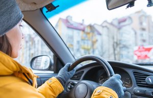 Eine Frau sitzt in einem Auto hinter dem Steuer. Sie trägt Winterkleidung mit einer gelben Daunenjacke, grauen Handschuhen und einer grauen Mütze.