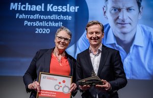 Michael Kessler hält eine Fahrrad-Bronzeskulptur in den Händen. Er lächelt in die Kamera. Gekleidet ist er mit einem dunkelblauen Anzug. Dazu trägt er ein weißes Hemd. Neben ihm auf der Bühne steht Christine Fuchs, Vorständin der Arbeitsgemeinschaft fußgänger-und fahrradfreundlicher Städte, Gemeinden und Kreise in NRW e.V. (AGFS). Sie hält die gerahmte Urkunde für die „Fahrradfreundlichste Persönlichkeit 2020“ in den Händen. Sie trägt einen dunkelblauen Hosenanzug und ein lachsfarbenes T-Shirt. Zudem hat sie eine Brille auf.