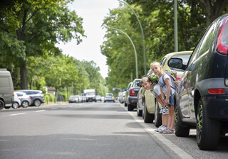 Ein Mädchen und ein Junge im Schulalter stehen zwischen parkenden Autos und schauen auf die Straße. 