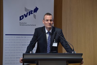 Auf dem Podium hinter dem Rednerpult steht Stefan Grieger. Er trägt einen schwarzen Anzug, ein weißes Hemd und eine Krawatte mit Blumenmuster. 
