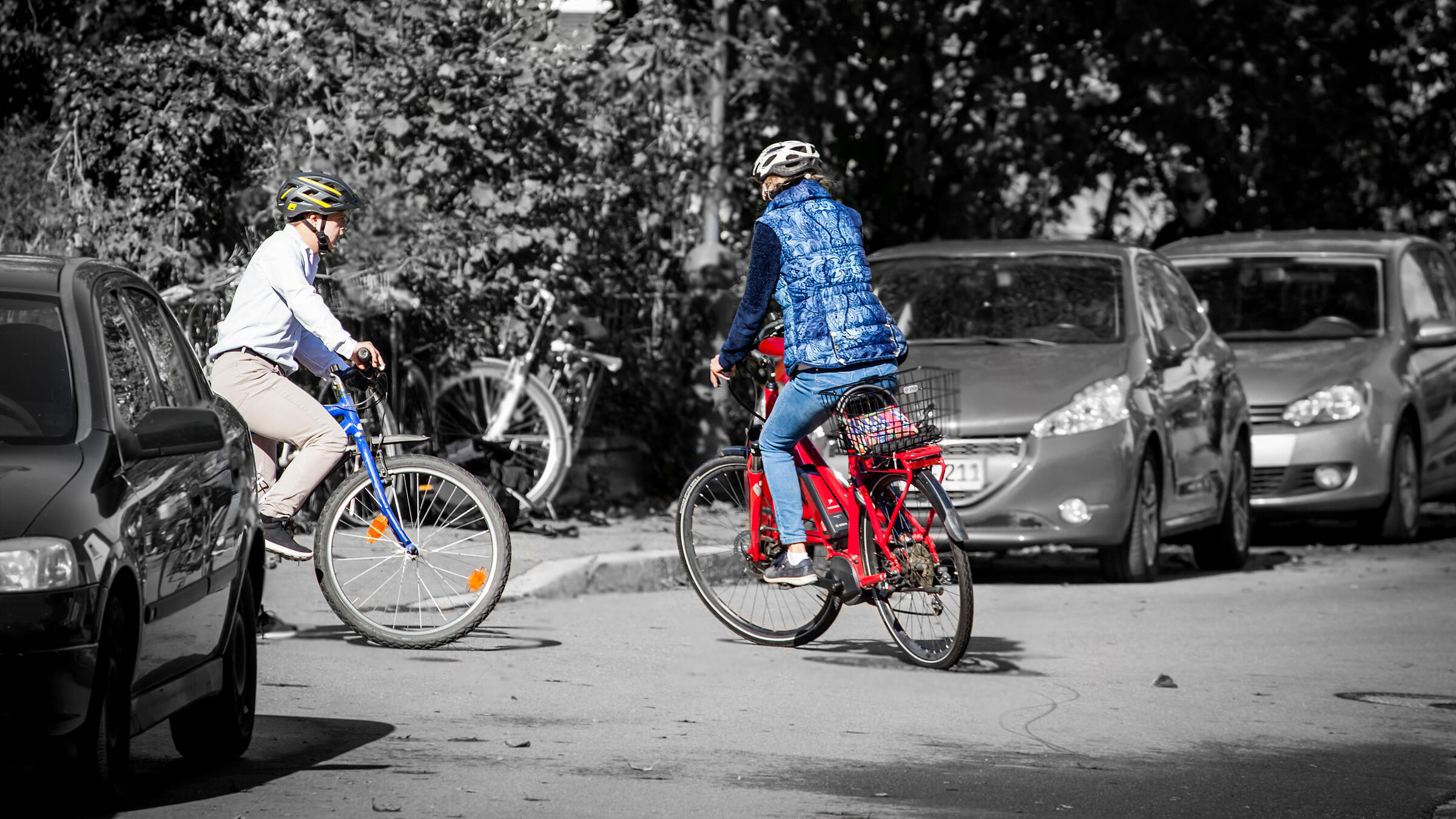 Zwei Fahrradfahrerinnen sind zu shene, die auf einen Weg abbiegen beziehungsweise von dem Weg auf die große Straße abbiegen. Sie tragen Helme.