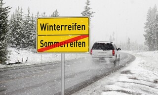 Ortsausgangsschild, oben steht Winterreifen, darunter, durchgestrichen Sommerreifen. Im Hintergrund fährt ein Auto durch eine Schneelandschaft