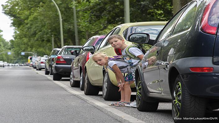 Ein Junge und ein Mädchen im Alter von sechs Jahren schauen zwischen parkenden Autos hervor.