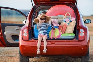 Der Kofferraum eines roten Kleinwagens ist geöffnet. Darin sind Koffer und Taschen für den Urlaub verstaut. Ein kleines Mädchen mit Sonnenhut und Brille sitzt links im Kofferraum und lächelt den Betrachter an. Foto: Adobe Stock