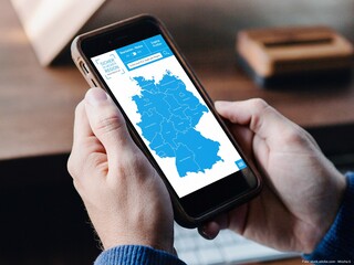 Zwei Männerhände halten ein Smartphone. Der Bildschirm zeigt eine Karte von Deutschland in blau.