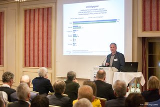 Jörg Ortlepp von der Unfallforschung der Versicherer (UDV) steht auf dem Podium hinter einem Rednerpult. Die Aufnahme ist aus der Perspektive des auf Stühlen sitzenden Publikums gemacht. Ein an die Wand projiziertes Bild zeigt einen Auszug aus der Unfallstatistik 2019.