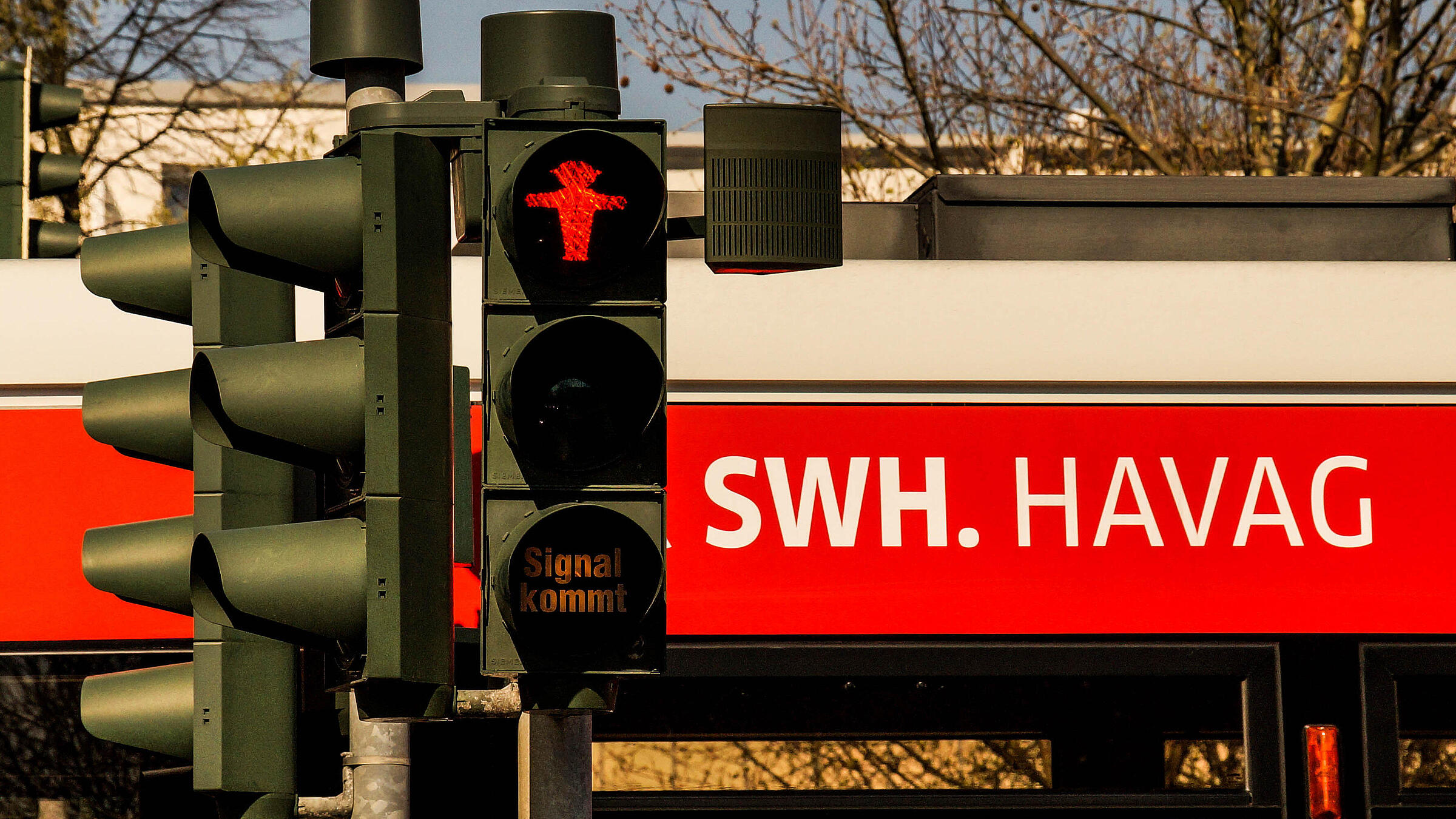 Eine Fußgängerampel zeigt ein rotes Halt-Symbol in Form einer stehenden Figur. Darunter befindet sich ein kleines Zusatzsignal mit der Anzeige „Signal kommt“. Im Hintergrund ist ein Haltestellenbereich mit dem Aufdruck „SWH. HAVAG“ zu sehen.