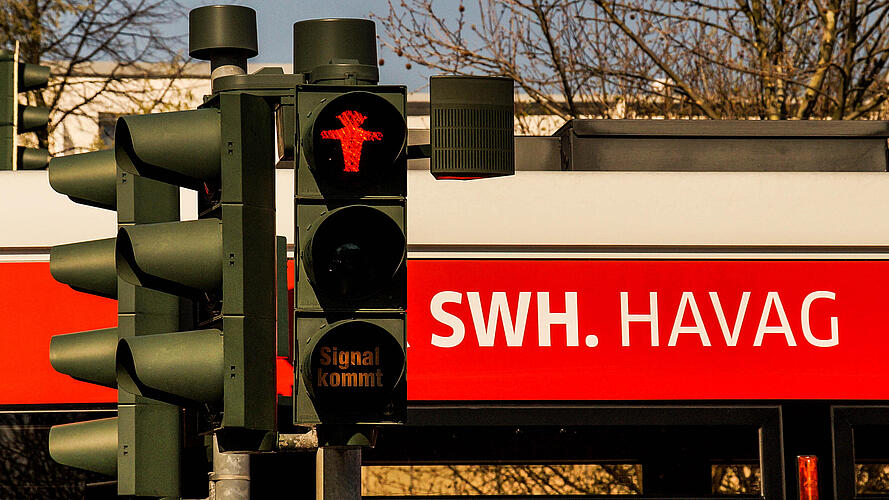 Eine Fußgängerampel zeigt ein rotes Halt-Symbol in Form einer stehenden Figur. Darunter befindet sich ein kleines Zusatzsignal mit der Anzeige „Signal kommt“. Im Hintergrund ist ein Haltestellenbereich mit dem Aufdruck „SWH. HAVAG“ zu sehen.
