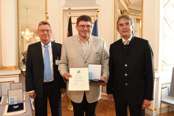 Bernhard Sigl, Foto: Tino Lex Drei Männer stehen nebeneinander. Der mittlere hält eine Urkunde