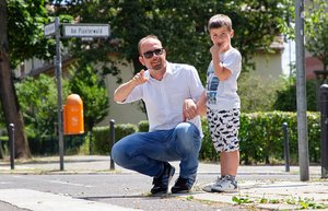 Ein Mann hockt neben einem kleinen Jungen. Er hält die Hand des Kindes. Beide schauen nach rechts in Richtung der Straße, die sie überqueren wollen. Es ist ein sonniger Tag.