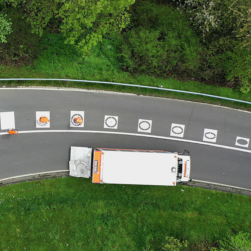 Menschen in orangener Arbeitskleidung bringen Fahrbahnmarkierungen in einer Kurve an. Die Markierungen sind Ellipsen in unterschiedlicher Größe.