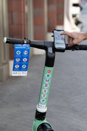 Eine Hand aktiviert einen E-Scooter per Handy, welches am Lenker des Tretrollers angebracht ist.