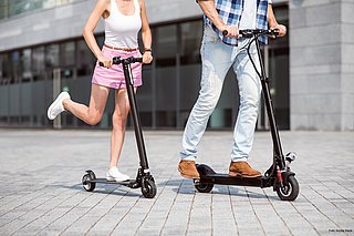 Eine Frau in kurzen rosa Hosen und mit einem weißen Tanktop sowie ein Mann in Jeans und Hemd rechts neben ihr, fahren auf grauen Pflastersteinen auf E-Scootern. Die Atmosphäre ist sommerlich.