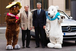 Links steht ein Mensch, verkleidet als Braunbär, daneben ein älterer Herr. Rechts von ihm steht ebenfalls ein Herr in dunklem Anzug, daneben ein Mensch, verkleidet als Eisbär. Im Hintergrund steht ein Auto.