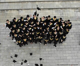 Viele junge Menschen stehen in einer Gruppe, tragen schwarze Umhänge und werden ihre schwarzen Universitätshüte in die Luft.