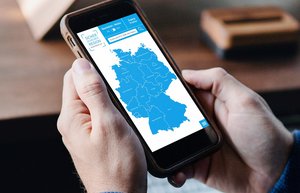 Zwei Männerhände halten ein Smartphone. Der Bildschirm zeigt eine Karte von Deutschland in blau.