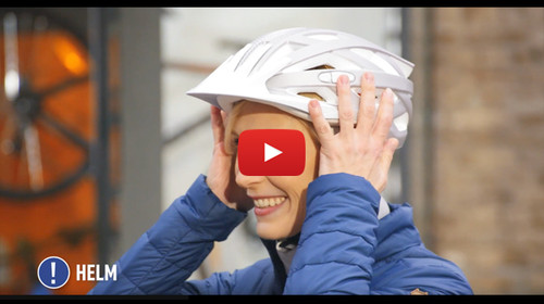 Vorschaubild für YouTube-Video „Sicher Pedelec fahren“: Eine Frau in blauer Jacke setzt einen weißen Fahrradhelm auf.