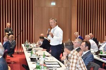 Ein Mann steht mit einem Mikrofon hinter einem Konferenztisch