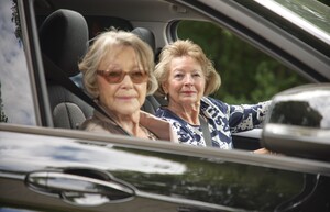 Zwei ältere Damen sitzen in einem schwarzen Auto. Eine von ihnen ist die Fahrerin des Pkws, die andere sitzt auf dem Beifahrersitz. Sie blicken in Richtung der Kamera, die ins Beifahrerfenster hinein fotografiert.