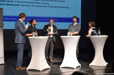 Die Fishbowl-Diskussion mit Fragen und Anmerkungen aus dem Publikum Vier Männer und eine Frau stehen im Halbkreis auf der Bühne. Vor ihnen sind drei weiße Stehtische platziert. Jeder von ihnen hat ein Handmikrofon. Der Mann in der Mitte spricht in seines und alle anderen blicken in seine Richtung.