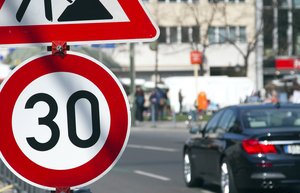 Am vorderen linken Bildrand ist ein Tempo 30-Schild. Dahinter fährt ein Auto entlang und Menschen stehen auf dem Gehweg. Der Hintergrund ist leicht verschwommen.