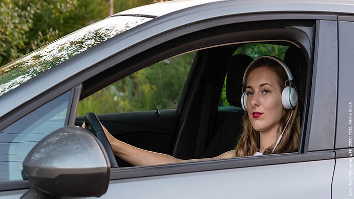 Eine Frau sitzt hinter dem Steuer eines Autos. Sie trägt weiße Kopfhörer auf den Ohren und blickt aus dem Autofenster.