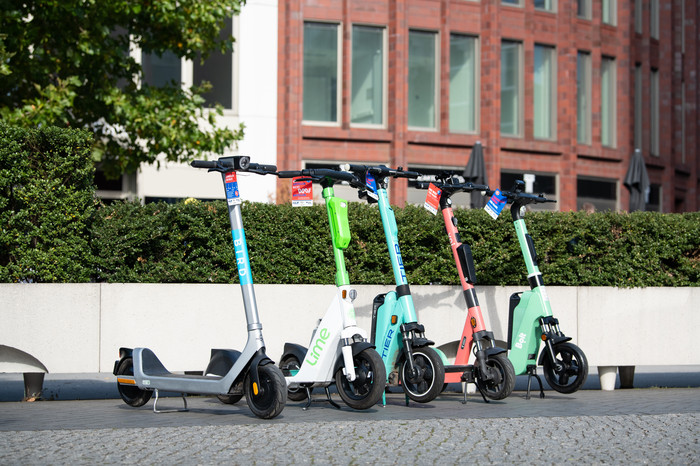 Fünf E-Scooter stehen ordentlich geparkt am Gehwegrand. Sie haben Flyer der Kampagne Roll ohne Risiko am Lenker.