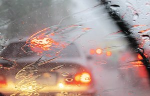 Durch die Windschutzscheibe eines Autos sind die Lichter eines vorausfahrenden Fahrzeugs zu erkennen. Auf der Windschutzscheibe sind Regentropfen, die mit leichten Schlieren von den Scheibenwischern zur Seite geschoben werden.