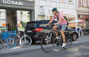 Ein Mann mit Helm, rosa Hemd und kurzen Hosen fährt mit einem Fahrrad entlang einer Straße mit Geschäften. Links im Bild ist ein Geschäft zu sehen, weitere abgestellte Fahrräder und ein schwarzes Auto.