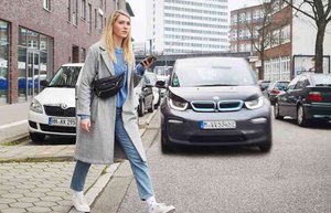 Es ist Herbst. Eine Frau im grauen Mantel und Jeans läuft über die Straße. Sie hält ein Handy in der Hand. Hinter ihr kommt ein Auto angefahren, wobei ihr Blick nicht in Richtung des Autos geht. Sie schaut vielmehr in die andere Richtung, ohne das Auto wahrzunehmen.