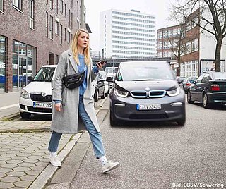 Es ist Herbst. Eine Frau im grauen Mantel und Jeans läuft über die Straße. Sie hält ein Handy in der Hand. Hinter ihr kommt ein Auto angefahren, wobei ihr Blick nicht in Richtung des Autos geht. Sie schaut vielmehr in die andere Richtung, ohne das Auto wahrzunehmen.