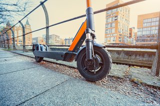 Ein orangefarbener E-Scooter steht am Gehwegrand auf einem Kiesabschnitt. Im Hintergrund stehen Häuser und Bäume ohne Blätter.