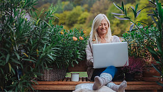 Older woman working from homeoffice, sitting on terrace with laptop.