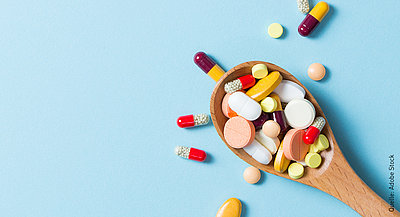 Viele verschieden geformte Tabletten in zahlreichen Farben, die auf einem Kochlöffel liegen. Manche der Tabletten liegen neben dem Kochlöffel auf einem hellblauen Untergrund. 