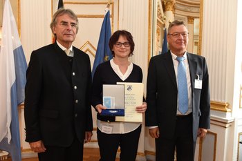 Hildegard Artner, Foto: Tino Lex Eine Frau steht zwischen zwei Männern und hält eine Urkunde