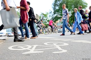 Fußgänger und Radfahrer überqueren bei Tageslicht und sommerlichen Temperaturen die Straße. Im Hintergrund warten Autos auf die Weiterfahrt.
