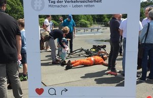 Im Vordergrund steht ein Rahmen aus Karton, auf dem Tag der Verkehrssicherheit steht. Blickt man durch den Rahmen sieht man einen Mensch in orangefarbenem Anzug auf dem Boden liegen. Um ihn herum stehen Menschen. Foto: DVR