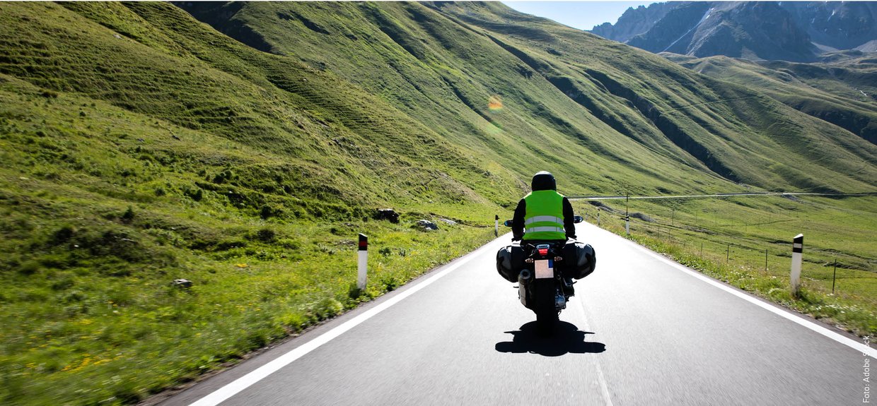 Ein Motorradfahrer fährt auf einer Landstraße durch die Berge. Die Sonne scheint.