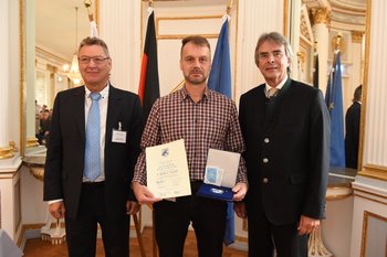 Christian Biersack, Foto: Tino Lex Drei Männer stehen nebeneinander. Der mittlere Mann hält eine Urkunde