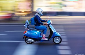 Eine Jugendliche fährt auf einem blauen Moped. Sie trägt einen silberfarbenen Helm und fährt auf einer Straße in der Dämmerung. Der Hintergrund ist verwischt.