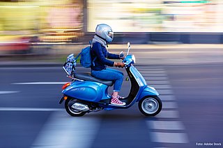 Eine Jugendliche fährt auf einem blauen Moped. Sie trägt einen silberfarbenen Helm und fährt auf einer Straße in der Dämmerung. Der Hintergrund ist verwischt.