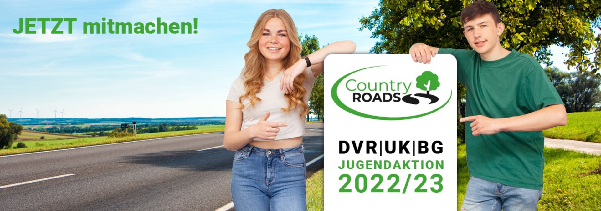 Zwei Jugendliche vor einem Banner "Country Roads" DVR/UK|BG 2022/23, im Hintergrund eine Landstraße im Sommer