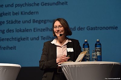 Der Impulsvortrag von Dr. Viktoria Wesslowski, Beraterin für kommunale Fußverkehrsförderung Eine Frau im Hosenanzug steht auf der Bühne. Sie trägt eine Brille und lächelt in Publikumsrichtung. Sie hat ein Kopfmikrofon und hält in der rechten Hand einen Presenter, um von einer PowerPoint-Folie zur nächsten zu wechseln. Mit dem linken Arm stützt sie sich leicht an einem Stehtisch ab, der auf der Bühne steht.
