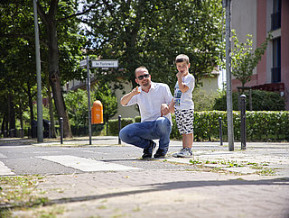 Ein Mann kniet neben einem Jungen an einem Zebrastreifen. Er zeigt nach rechts, der Junge blickt nach rechts.