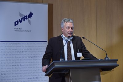Prof. Dr. Dieter Müller steht auf dem Podium hinter dem Rednerpult. Er trägt einen schwarzen Anzug, ein weißes Hemd und eine braun karierte Krawatte.
